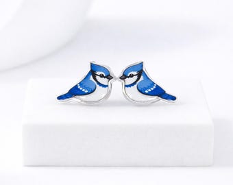 May include: A pair of blue jay stud earrings. Each earring features a blue and white bird design with a silver-colored outline. The birds face each other, set against a white background.