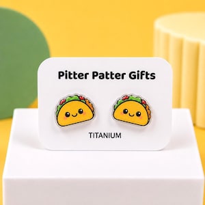 May include: A pair of stud earrings shaped like smiling tacos with yellow shells, green lettuce, and red toppings. The earrings are displayed on a white rectangular card labeled Pitter Patter Gifts and TITANIUM in black text.