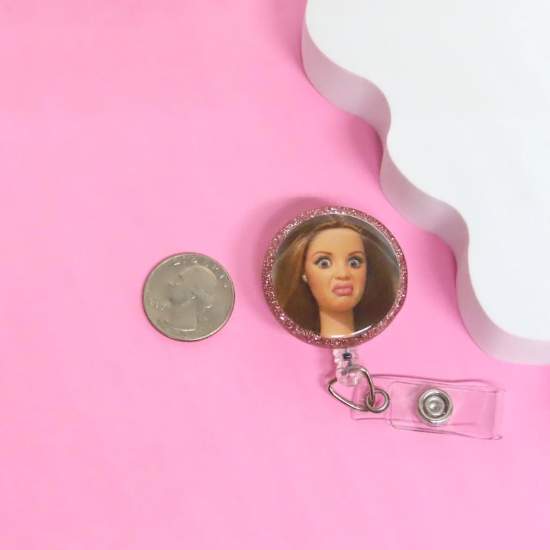 Puede incluir: Un portatarjetas redondo con un marco rosa brillante, que presenta la cabeza de una mu&ntilde;eca con una expresi&oacute;n de sorpresa. Tiene una correa de pl&aacute;stico transparente y un clip de metal. Una moneda est&aacute; al lado sobre una superficie rosa.