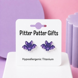 May include: A pair of purple bat-shaped stud earrings displayed on a white card labeled Pitter Patter Gifts and Hypoallergenic Titanium. The earrings feature a cartoon bat design with small wings, pointed ears, and a smiling face.