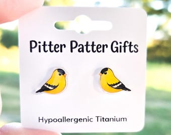 May include: A pair of stud earrings shaped like yellow goldfinches with black wing and head details. The earrings are mounted on a white display card labeled Pitter Patter Gifts and Hypoallergenic Titanium in black text.