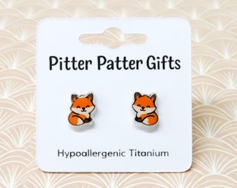 May include: A pair of stud earrings shaped like orange and white foxes on a white display card. The card features the text Pitter Patter Gifts at the top and Hypoallergenic Titanium at the bottom, indicating the material of the earring posts.