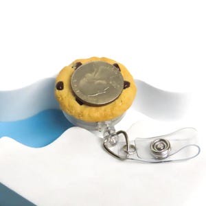 May include: A novelty cookie-shaped retractable badge holder with a quarter coin on top. The badge reel is light brown with chocolate chips and a clear plastic strap and metal clip.