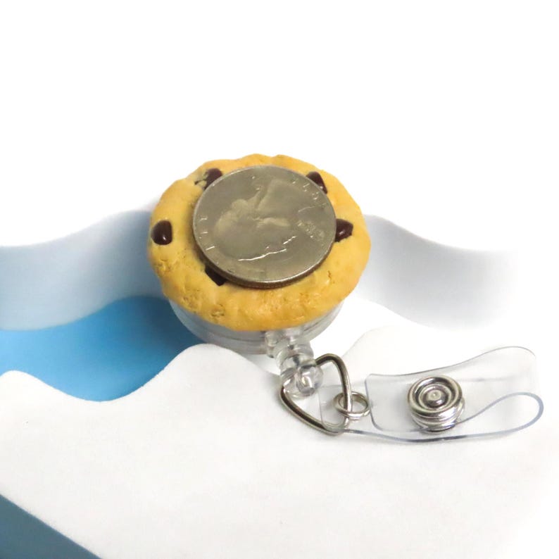 May include: A novelty cookie-shaped retractable badge holder with a quarter coin on top. The badge reel is light brown with chocolate chips and a clear plastic strap and metal clip.