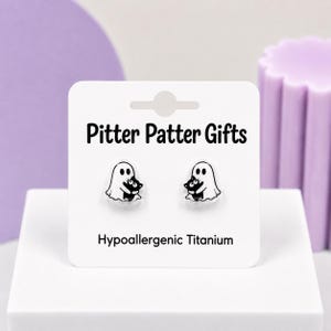 May include: A pair of stud earrings featuring a white ghost holding a black cat, displayed on a white card labeled Pitter Patter Gifts and Hypoallergenic Titanium. The earrings are made of a clear material with black and white graphic details.