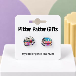 May include: A pair of stud earrings on a white display card labeled Pitter Patter Gifts and Hypoallergenic Titanium. One earring shows an open book on a stack, the other a stack of three closed books with a flower and heart graphic.