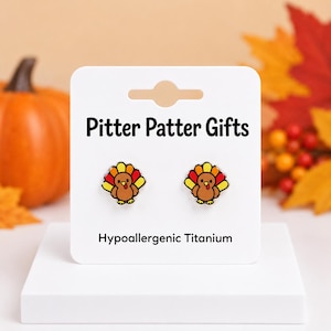 May include: A pair of stud earrings featuring a cartoon turkey design with brown, red, orange, and yellow feathers. The earrings are attached to a white display card labeled Pitter Patter Gifts and Hypoallergenic Titanium in black text.