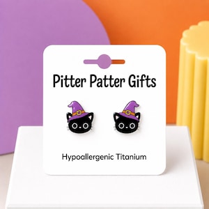 May include: A pair of black cat stud earrings featuring purple witch hats with orange bands. The earrings are displayed on a white card labeled Pitter Patter Gifts and Hypoallergenic Titanium in black text.