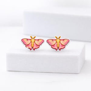 May include: A pair of moth-shaped stud earrings. The earrings feature a pink and yellow color scheme with a silver outline. The moth's wings are a gradient of pink and yellow, with a yellow body and antennae. The earrings are displayed on a white surface.