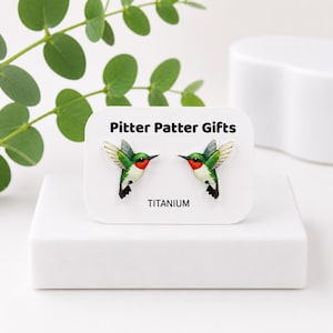 May include: A pair of stud earrings shaped like hummingbirds with green, white, and red plumage. The earrings are displayed on a white card labeled Pitter Patter Gifts and TITANIUM. The birds are depicted in flight with wings extended outward.