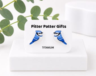 Puede incluir: Un par de pendientes de botón con forma de arrendajos azules con plumaje blanco y azul, picos negros y marcas negras en los ojos. Los pendientes están montados en una tarjeta blanca con el texto Pitter Patter Gifts y TITANIUM.