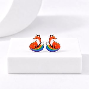 May include: A pair of stud earrings featuring a stylized fox illustration. Each fox has orange fur with white accents on the chest and face, and a tail that transitions into a vibrant rainbow gradient of red, orange, yellow, green, blue, and violet.