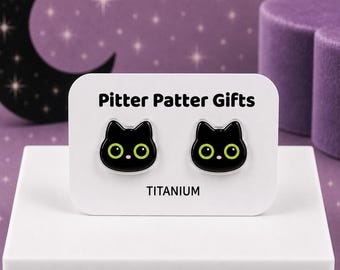 May include: A pair of stud earrings shaped like black cat heads with large lime green eyes and small pink noses. The earrings are mounted on a white display card labeled Pitter Patter Gifts and TITANIUM in black text, featuring a cat-ear silhouette.