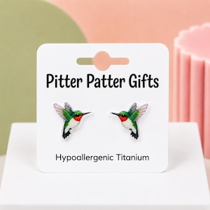 May include: A pair of stud earrings featuring a colorful hummingbird design with green, white, and red plumage. The earrings are displayed on a white card labeled Pitter Patter Gifts and Hypoallergenic Titanium, showing the bird in flight.