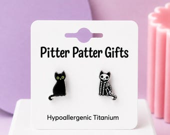 May include: A pair of stud earrings on a white display card labeled Pitter Patter Gifts and Hypoallergenic Titanium. One earring is a black cat with yellow eyes, and the other is a black cat with a white skeletal bone design.