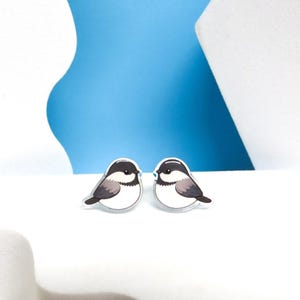 May include: A pair of bird-shaped earrings. Each earring features a black and white bird design with gray shading. The earrings are set against a white surface with a blue and white backdrop.