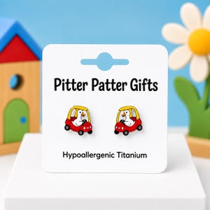 May include: A pair of stud earrings on a white display card labeled Pitter Patter Gifts and Hypoallergenic Titanium. The earrings feature a white duck with an orange beak driving a small red car with a yellow roof and black wheels.