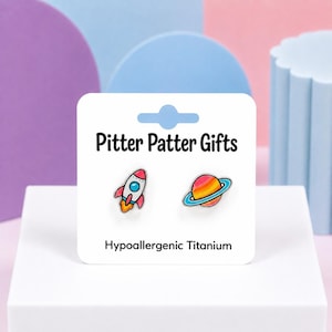 May include: A pair of hypoallergenic titanium stud earrings featuring a colorful rocket ship and a planet with rings. The earrings are displayed on a white card labeled Pitter Patter Gifts and Hypoallergenic Titanium in black text.