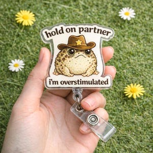 May include: A clear badge reel with a cartoon frog wearing a cowboy hat. The badge has the text "hold on partner" and "I'm overstimulated". The reel is held in a hand, with daisies and yellow flowers in the background.
