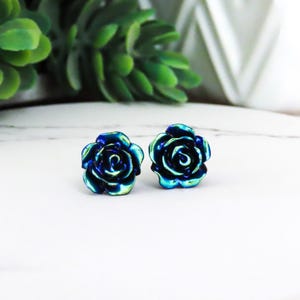 May include: A pair of iridescent blue rose-shaped stud earrings. The earrings have a metallic finish that reflects shades of blue and green. The floral design is detailed and realistic, perfect for adding a touch of nature-inspired elegance to any outfit.