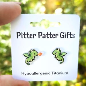 May include: A pair of green dragon stud earrings on a white display card labeled Pitter Patter Gifts and Hypoallergenic Titanium. One dragon is breathing a small orange flame, while the other is smiling. The earrings are made of titanium.