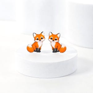 May include: A pair of stud earrings featuring a cartoon fox design. Each fox is orange with a white chest, face, and tail tip, and black paws. The foxes have large black eyes and a small black nose, rendered in a flat, illustrated style.