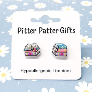 May include: A pair of stud earrings on a white display card labeled Pitter Patter Gifts and Hypoallergenic Titanium. One earring features an open book, and the other shows a stack of three colorful books with heart and flower details.