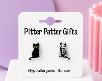 May include: A pair of stud earrings on a white display card labeled Pitter Patter Gifts and Hypoallergenic Titanium. One earring is a black cat with yellow eyes, and the other is a black cat with a white skeletal bone print design.