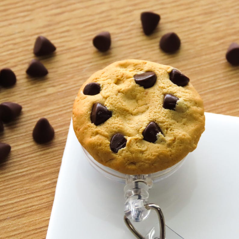 May include: A novelty retractable badge holder featuring a detailed chocolate chip cookie design. The cookie is a light brown color with dark chocolate chips. The badge reel has a clear plastic casing and a metal clip.