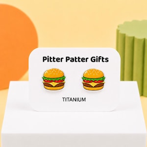 May include: A pair of stud earrings featuring a cartoon hamburger design with a golden bun, green lettuce, red tomato, yellow cheese, and a brown patty. The earrings are displayed on a white card labeled Pitter Patter Gifts and TITANIUM.