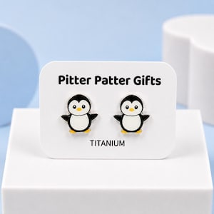 May include: A pair of titanium stud earrings shaped like penguins with black, white, and yellow enamel details. The earrings are attached to a white display card featuring the text Pitter Patter Gifts and the word TITANIUM printed below them.