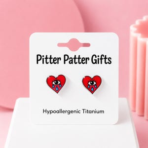 May include: A pair of heart-shaped stud earrings on a white display card labeled Pitter Patter Gifts and Hypoallergenic Titanium. Each red heart features a central eye with blue teardrops, rendered in a graphic style with black outlines.