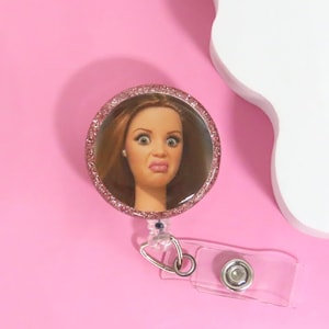 May include: A round, pink glitter badge reel with a clear plastic strap. The badge reel features a circular image of a woman with a surprised expression. The background is a solid pink.