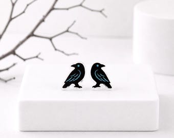 May include: A pair of stud earrings featuring a black raven design with light blue wing accents and a small light blue eye detail. The birds are rendered in a flat graphic style with clear outlines, positioned side by side on a white surface.