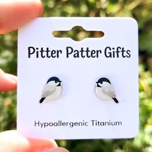 May include: A pair of stud earrings featuring a black, white, and gray chickadee bird design on a white display card. The card is printed with the text Pitter Patter Gifts and Hypoallergenic Titanium in black sans-serif font.