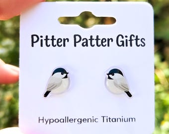 May include: A pair of stud earrings featuring a black, white, and gray chickadee bird design on a white display card. The card is printed with the text Pitter Patter Gifts and Hypoallergenic Titanium in black sans-serif font.