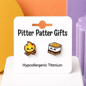 May include: A pair of enamel stud earrings on a white display card labeled Pitter Patter Gifts and Hypoallergenic Titanium. One earring is a smiling campfire, and the other is a smiling s'more with a graham cracker, marshmallow, and chocolate.