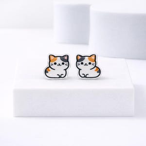 May include: A pair of cat-shaped stud earrings. Each earring features a white cat with black, orange, and brown markings. The earrings are set against a white background.