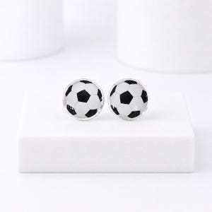 May include: A pair of round stud earrings featuring a soccer ball design. Each earring has a black and white pattern, resembling a soccer ball. The earrings are set against a white background.