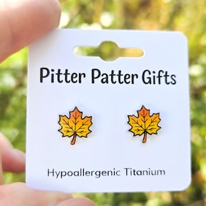 May include: A pair of stud earrings shaped like maple leaves with a yellow to orange gradient, mounted on a white display card labeled Pitter Patter Gifts and Hypoallergenic Titanium.