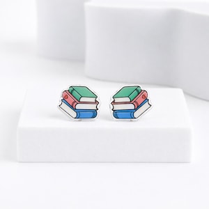 May include: A pair of book-shaped earrings. Each earring features a stack of three books in blue, red, and green, with a small heart detail on the red book. The earrings are set against a white background.