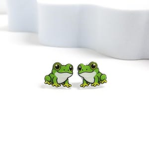 May include: A pair of stud earrings featuring a cartoon frog design. Each earring displays a bright green frog with large black eyes, a white belly, and yellow-tipped toes, rendered in a flat, graphic style with a clear, glossy finish.