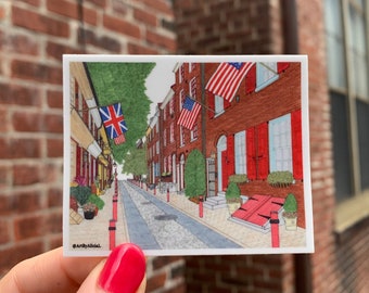 May include: A hand-drawn illustration of a narrow street in a city with red brick buildings. American and British flags hang from the buildings. The street is lined with trees and lampposts. The illustration is framed in a white border with the text "@ArtByAlicia" in the bottom left corner.