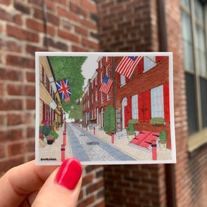 May include: A hand-drawn illustration of a narrow street in a city with red brick buildings. American and British flags hang from the buildings. The street is lined with trees and lampposts. The illustration is framed in a white border with the text "@ArtByAlicia" in the bottom left corner.