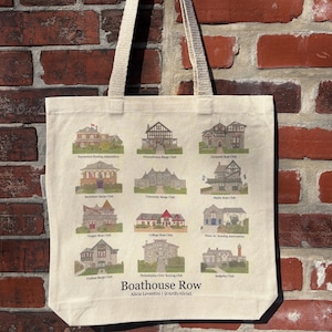 May include: A cream-colored tote bag with a print of Boathouse Row buildings. The bag has beige handles and is hanging against a red brick wall. The text "Boathouse Row" and the artist's name are printed on the bag.