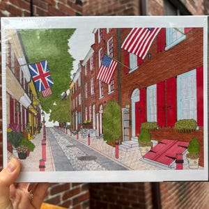 May include: A rectangular jigsaw puzzle box featuring a detailed illustration of a historic brick street scene. The image displays red brick buildings with red shutters, green potted plants, and American flags hanging from the building facades.