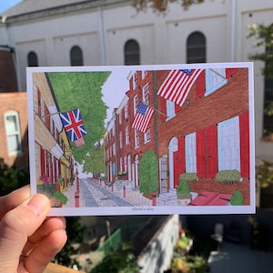 May include: A postcard illustration of Elfreth's Alley in Philadelphia, Pennsylvania. The alley is lined with brick buildings with red shutters and American flags hanging from the windows. The alley is paved with cobblestones and there are trees and plants growing along the sides.