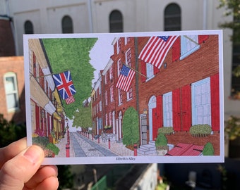 May include: A postcard illustration of Elfreth's Alley in Philadelphia, Pennsylvania. The alley is lined with brick buildings with red shutters and American flags hanging from the windows. The alley is paved with cobblestones and there are trees and plants growing along the sides.