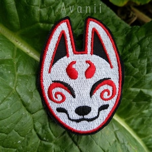 May include: White embroidered fox mask with red eyes and a black nose. The mask is surrounded by a red border and is on a green leaf.