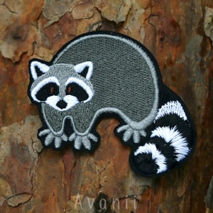 Op de afbeelding: Geborduurde strijkpatch van een grijze wasbeer met een wit gezicht en zwarte ogen. De wasbeer heeft een wit-zwarte gestreepte staart.